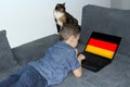 German flag on a laptop display, a little schoolboy in jeans lies on a sofa and scrolls, a cat sits nearby, a concept of learning