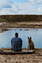 German shepherd with owner. Rear view. Young man with dreadlocks is sitting on wooden log on riverbank with dog enjoying views of Royalty Free Stock Photo