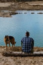 German shepherd with owner. Rear view. Young man with dreadlocks is sitting on wooden log on riverbank with dog enjoying views of Royalty Free Stock Photo