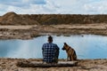 German shepherd with owner. Rear view. Young man with dreadlocks is sitting on wooden log on riverbank with dog enjoying views of Royalty Free Stock Photo