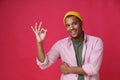 Gesturing ok, okay smiling african american young man wearing pink shirt and yellow hat isolated on red background. Happy trendy Royalty Free Stock Photo