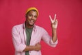 Gesturing V, victory smiling african american young man wearing pink shirt and yellow hat isolated on red background Royalty Free Stock Photo