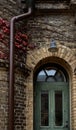 Ghastly green arched door on bricked disquiet facade with red ivy leaves and a water spout beside conveys restlessness