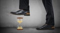 Giant male legs in shiny boots stand on concrete background and step on a small hourglass.