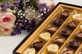 Bouquet of pink roses with baby breath and purple flowers next to fancy box of chocolates.