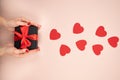 Gift box wrapped in black paper with red ribbon in female hand on pink surface. Flying red hearts symbol of love Royalty Free Stock Photo