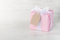 Gift box wrapped in pink dotted paper, tied satin bow and an empty kraft card over a white wood background.