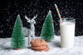 A glass of milk with a straw and cookies with chocolate chips on a snowy background among toy Christmas trees Royalty Free Stock Photo