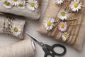 Gifts packed in burlap fabric with beautiful chamomiles, ropes, spool of threads and scissors on white wooden table, flat lay