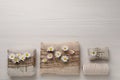 Gifts packed in burlap fabric with beautiful chamomiles, ropes and spool of threads on white wooden table, flat lay. Space for