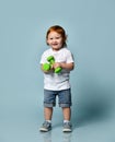Ginger baby boy or girl in white t-shirt, shoes, denim shorts. Smiling, holding two green dumbbells, posing on blue background Royalty Free Stock Photo