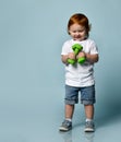 Ginger baby boy or girl in white t-shirt, shoes, denim shorts. Smiling, holding two green dumbbells, posing on blue background Royalty Free Stock Photo