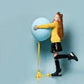 Teenage kid in yellow sweatshirt, black skirt, knee-highs, boots. Smiling, holding balloon, posing sideways on blue background.