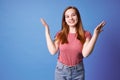 Ginger girl in pink t-shirt and jeans posing  on blue. Girl raised her hands, admiring gesture. Royalty Free Stock Photo