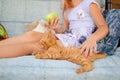 Ginger kitten lies near the girl on the garden swing. The girl strokes her pet. Little blonde caucasian girl with a red cat Royalty Free Stock Photo