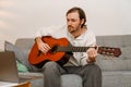 Ginger man playing guitar and using laptop while sitting on couch Royalty Free Stock Photo