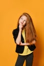 Ginger teenage child in black jacket and pants, yellow t-shirt. She is looking thoughtful, posing on orange background. Close up