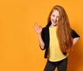 Ginger teenage girl in black jacket, pants, yellow t-shirt. She smiling, showing okay sign, posing on orange background. Close up