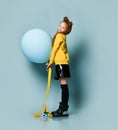 Ginger teenage girl in yellow sweatshirt, black skirt, knee-highs, boots. She smiling, holding balloon, posing on blue background.