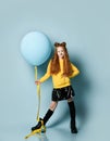 Ginger teenage girl in yellow sweatshirt, black skirt, knee-highs, boots. She smiling, holding balloon, posing on blue background.