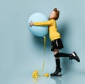 Teenage kid in yellow sweatshirt, black skirt, knee-highs, boots. Smiling, holding balloon, posing sideways on blue background.