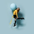 Teenager in yellow sweatshirt, black skirt, knee-highs, boots. She smiling, holding balloon, jumping up against blue background.