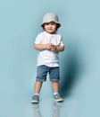 Ginger toddler boy or girl in white t-shirt, gray hat, socks and shoes, denim shorts. Child is smiling, posing on blue background Royalty Free Stock Photo