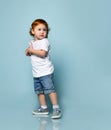 Ginger toddler boy or girl in white t-shirt, socks and shoes, denim shorts. Holding sunglasses, posing sideways on blue background Royalty Free Stock Photo