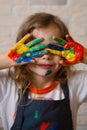 A girl artist in a creative workshop covers her face with her hands stained with paint on the background of a brick wall.