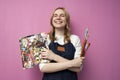 Girl artist holds brushes and a palette and smiles on a pink background, student of art school, profession of an artist