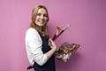 Girl artist holds brushes and a palette and smiles on a pink background, student of art school, profession of an artist