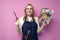 Girl artist holds brushes and a palette and smiles on a pink background, student of art school, profession of an artist