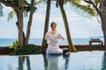 Woman from the back is sitting in lotus pose, stretching hands up, meditating on pool on the coast of ocean in Indonesia Royalty Free Stock Photo