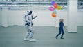 A girl with balloons is dancing and a manlike robot is coming towards her