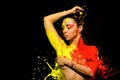 A girl is bathed in yellow paint on one side and orange paint on the other. With one hand, the girl closes her breast, and t