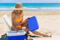 girl in bathing suit and hat sits on chair on the beach and takes out cold drinks from refrigerator Royalty Free Stock Photo