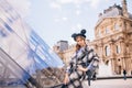 A girl in a beautiful coat and Mickey mouse hat sits on a bench near the Louvre