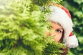 Girl with beautiful eyes in a santa claus hat hiding behind a christmas tree. Concept. Royalty Free Stock Photo