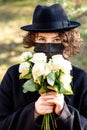 Girl in a black coat, hat and black disposable mask with a bouquet of white roses outdoors. Sunny spring day