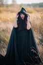 A girl in a black dress, a cloak with a hood stands in a high dry grass in the field against the background of the forest. Witch C