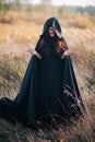 A girl in a black dress, a cloak with a hood stands in a high dry grass in the field against the background of the forest. Witch C