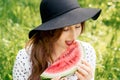 A girl in black hat is eating piece of watermelon on green background.