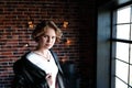 The girl in a black jacket and a light blouse poses opposite to a window, a background the brick wall covered with glow lamps. Royalty Free Stock Photo