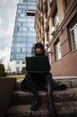 Girl in black leather coat and hood with laptop on city street, concept of woman hacker and agent working remotely at computer Royalty Free Stock Photo