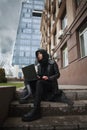 Girl in black leather coat and hood with laptop on city street, concept of woman hacker and agent working remotely at computer Royalty Free Stock Photo