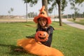 Girl in black shirt, witch hat and orange skirt offering pumpkin to receive candy and sitting on grass celebrating halloween.
