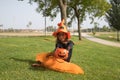 Girl in black shirt, witch hat and orange skirt offering pumpkin to receive candy and sitting on grass celebrating halloween.
