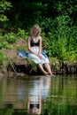 A girl with blond hair on the lake shore has her feet in the water and is swinging them creating a spray of water. Royalty Free Stock Photo