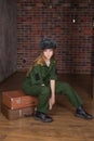 A girl with blond hair in military uniform and a winter hat sits on suitcases against a brick wall. February 23. portrait of a mil