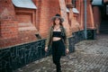 girl with blond long hair, in black leggings, a tank top, a beige jacket and a hat, walking on the street Royalty Free Stock Photo